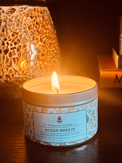 A lit Ocean Breeze scented candle, a 100ml tin from The Victorian Candle Co. made with vegan-friendly soy wax, is placed on a dark surface. It casts a warm glow beside a decorative, mosaic-patterned candle holder.