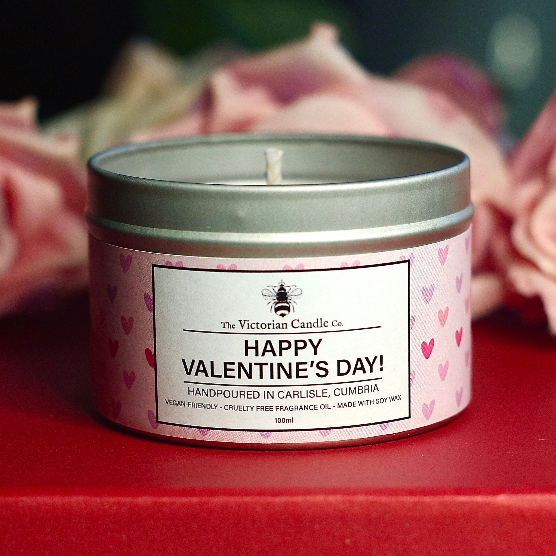 The Victorian Candle Co.s 100ml Pink Fizz scented candle features a pink heart pattern label reading Happy Valentines Day! It is crafted with soy vegan-friendly wax and placed on a red surface, with blurred pink roses in the background. This is a Valentines Day Exclusive.