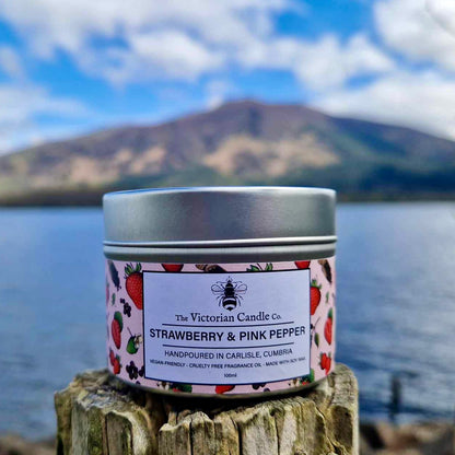 A Strawberry & Pink Pepper scented candle from The Victorian Candle Co., packaged in a 100ml tin, fills the air with a delicate soft musk aroma while resting on a weathered wooden post. In the background, a tranquil lake and mountain gently fade into the scene, enhancing the candle's serene ambiance.