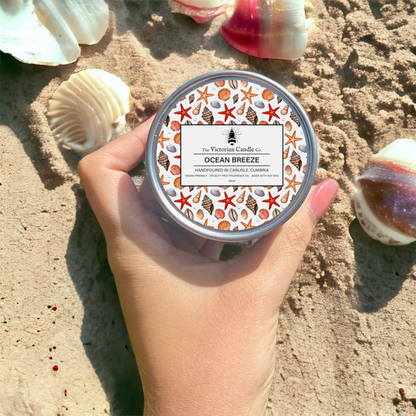 A hand holds The Victorian Candle Co.'s Ocean Breeze - Scented Candle in a 100ml tin, surrounded by seashells on sandy ground. This vegan-friendly soy wax candle features a tin adorned with starfish and shell patterns, emitting a delightful fruity floral scent.