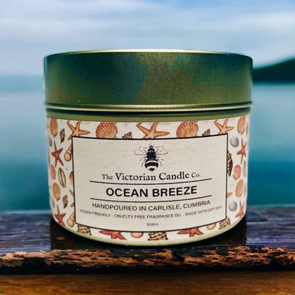 A green tin candle called Ocean Breeze - Scented Candle - 100ml Tin from The Victorian Candle Co., adorned with illustrations of seashells and starfish, is crafted using vegan-friendly wax and soy wax. Handmade in Carlisle, Cumbria, its label highlights a fruity floral scent against a blurred ocean and sky backdrop.