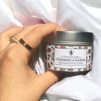 A hand holds a 100ml tin Strawberry & Rhubarb scented candle by The Victorian Candle Co., set against white fabric with two rings, featuring charming strawberry designs on its label.