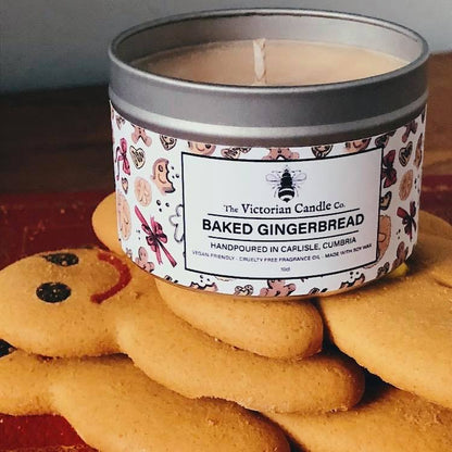 A tin of Baked Gingerbread - Scented Candle - 100ml Tin 🍪 by The Victorian Candle Co. is placed on gingerbread cookies. The candle's label showcases festive designs, and the cookies are shaped like gingerbread people with smiling faces.