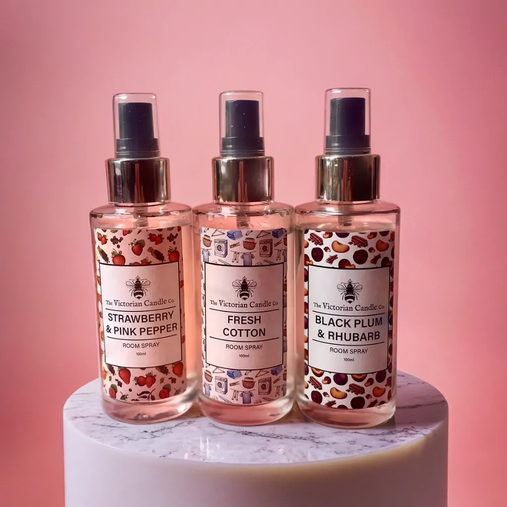 The Room Spray Trio Deal from The Victorian Candle Co features three captivating room sprays elegantly arranged against a pink backdrop on a white circular stand. The enticing scents offered are Strawberry & Pink Pepper, Fresh Cotton, and Black Plum & Rhubarb, each adorned with vibrant patterns on their labels.