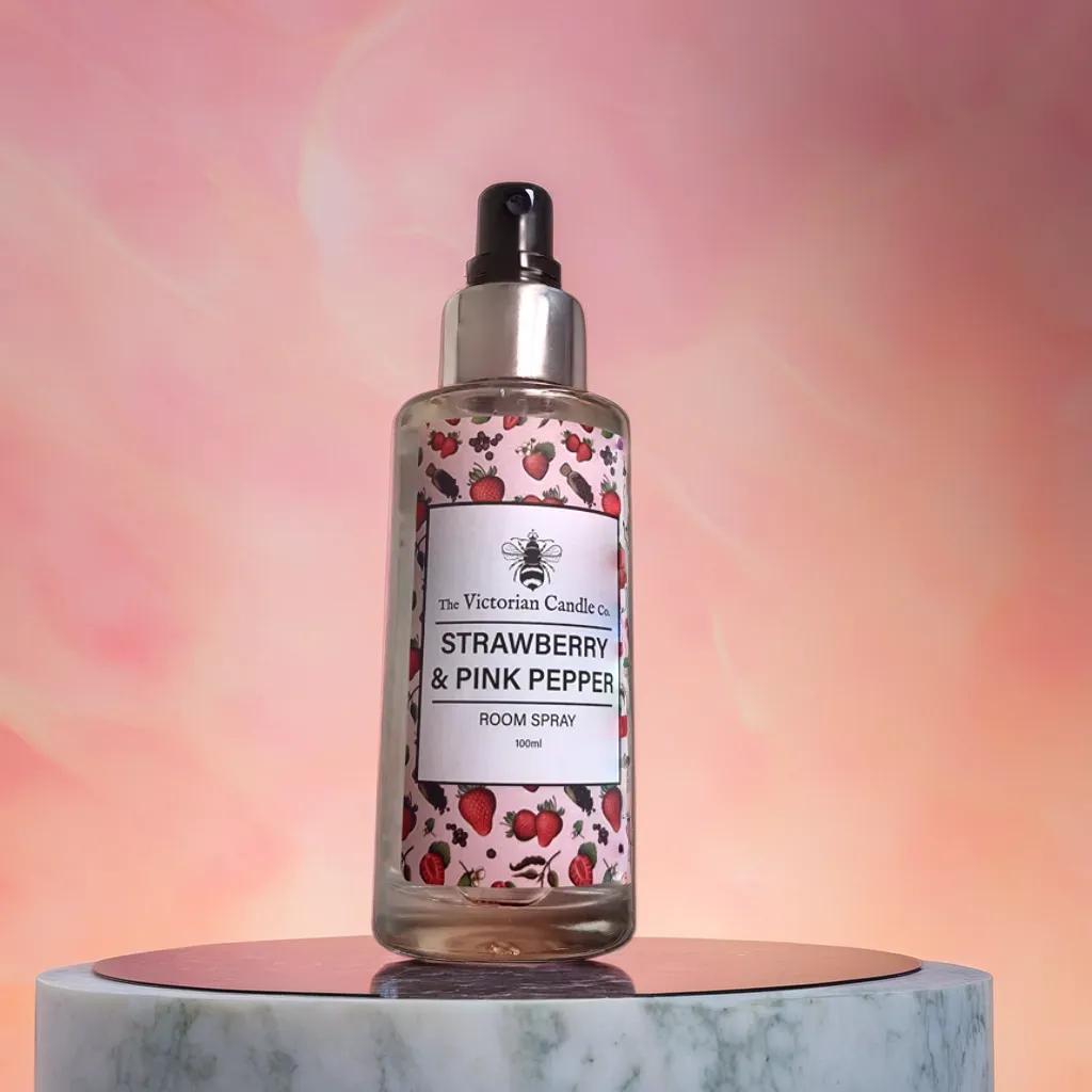 The Strawberry & Pink Pepper - Room Spray by The Victorian Candle Co is elegantly placed on a reflective surface, set against a stunning pink and orange gradient backdrop. Its label, adorned with delightful images of strawberries and pink peppercorns, evokes captivating fragrances.