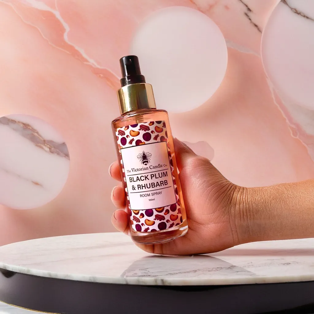 A hand holds a bottle of The Victorian Candle Co's elegant Black Plum & Rhubarb Room Spray, showcasing a fruit-themed label and a sophisticated black spray top, beautifully positioned against a pink and marble backdrop on a round marble surface.