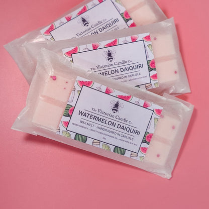 On a pink surface, three Watermelon Daiquiri Wax Melt Snap Bars from The Victorian Candle Co. are displayed. Each bar showcases a decorative label adorned with charming watermelon illustrations. The light pink wax melts are sprinkled with small pink dots, adding to their delightful appearance.