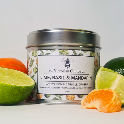 A large, 200ml tin of The Victorian Candle Co.'s Lime, Basil & Mandarin scented candle is elegantly displayed against a backdrop featuring vibrant illustrations of limes, basil, and mandarins. Surrounded by fresh lime pieces, mandarins, and basil leaves, the candle proudly showcases its vegan-friendly and cruelty-free commitment.