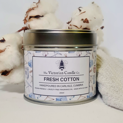 A 200ml Fresh Cotton Scented Candle in a recyclable tin from The Victorian Candle Co. rests beside cotton flowers and a knitted cloth. This hand-poured candle, crafted in Carlisle, Cumbria, is made with soy vegan wax.