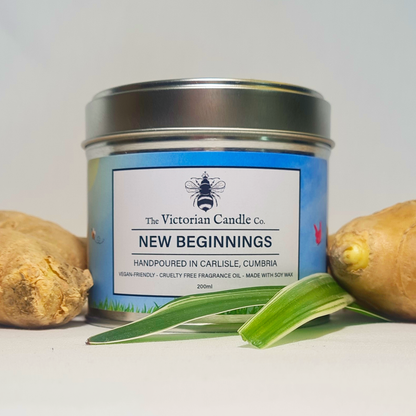 A large 200ml tin candle named New Beginnings by The Victorian Candle Co. sits between two pieces of ginger, filling the air with a refreshing lemongrass and ginger scent. It features vegan-friendly, cruelty-free fragrance oil and soy wax, beautifully set against a grassy background.