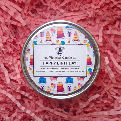 A 100ml tin scented candle called Happy Birthday from The Victorian Candle Co. is nestled atop pink shredded paper. The label is adorned with cheerful illustrations of cakes, balloons, and gifts, making it perfect for a celebratory occasion. This vegan-friendly candle is crafted with soy wax for a delightful birthday experience.