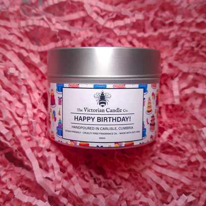 A Happy Birthday - Scented Candle - 100ml Tin from The Victorian Candle Co. rests on pink shredded paper, adorned with vibrant birthday-themed illustrations on its label. This handmade soy wax candle crafted in Carlisle, Cumbria, is perfect for celebrating with style.