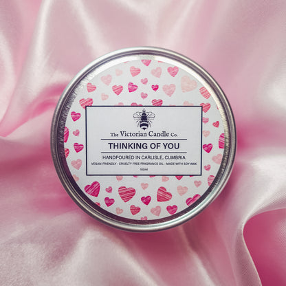 A round tin candle decorated with pink and red heart designs on the lid, labeled as "The Victorian Candle Co. Thinking of You." Handcrafted in Carlisle, Cumbria, using soy vegan-friendly wax and cruelty-free fragrance oil, it is showcased on pink satin fabric.