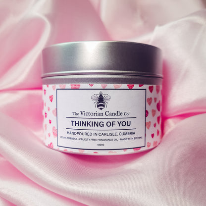 A pink candle tin from The Victorian Candle Co. labeled "Thinking of You - Scented Candle - 100ml Tin" rests on a soft pink fabric background. Made with soy vegan-friendly wax, the label indicates it is hand-poured in Carlisle, Cumbria, and is cruelty-free.