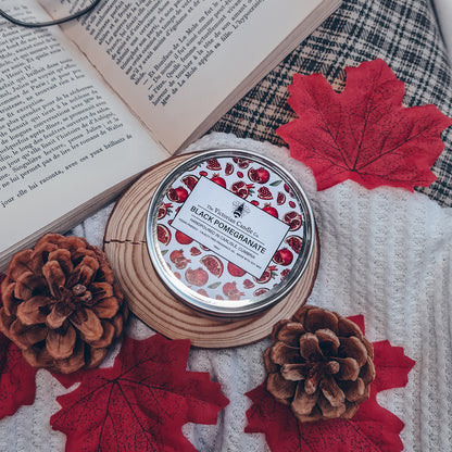 A tin of Black Pomegranate - Scented Candle by The Victorian Candle Co., crafted from soy wax and housed in a 100ml tin, rests on a wooden coaster, surrounded by pinecones and red maple leaves. An open book with beige pages lies beside the tin on a textured white and plaid surface, creating an inviting autumn scene.