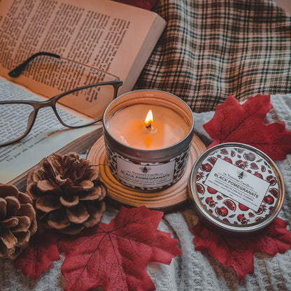 A 100ml tin of The Victorian Candle Co.'s Black Pomegranate scented candle sits on a coaster, surrounded by red autumn leaves, pinecones, and an open book. Glasses rest on the book, while a checked fabric is partially visible in the background, enhancing this cozy and vegan-friendly setup.