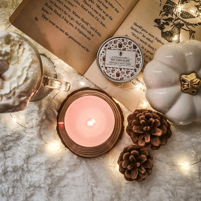 An inviting scene features an open book, a cup of vegan-friendly hot chocolate topped with whipped cream, the warm glow of The Victorian Candle Co.'s Hot Chocolate - Scented Candle in a 100ml tin lit nearby, alongside pinecones, fairy lights, and a decorative pumpkin. The ambiance is warm and perfect for a relaxing autumn evening.