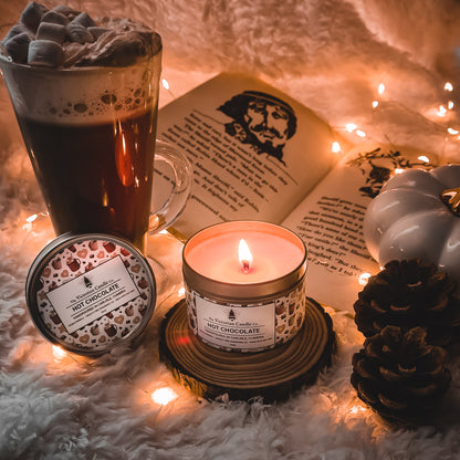 A cozy scene unfolds with a lit 100ml tin of The Victorian Candle Co.'s Hot Chocolate scented candle, an open book, and a mug of hot chocolate topped with marshmallows. Twinkling fairy lights add a warm glow, while a white pumpkin and pinecone enhance the autumnal atmosphere in this vegan-friendly nook.