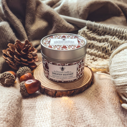 A cozy scene showcases a Sweater Weather scented candle from The Victorian Candle Co., crafted with soy vegan-friendly wax, flickering on a small wooden platform. Surrounded by acorns, pine cones, and thick knit blankets, it creates a warm, autumnal atmosphere with hints of citrus grapefruit.