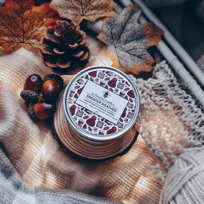 A cozy autumn-themed scene showcases The Victorian Candle Co.'s Sweater Weather scented candle in a 100ml tin, crafted from soy vegan-friendly wax. It is surrounded by knitted fabric, autumn leaves, a pine cone, and acorns, with the warm lighting enhancing the fall atmosphere.