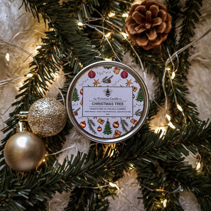 A lit Christmas Tree - Scented Candle in a 100ml tin from The Victorian Candle Co., crafted from soy wax, sits amidst a bed of green pine branches and is encircled by warm fairy lights. The festive arrangement is completed with two decorative balls—one gold and one glittery—and a brown pinecone.