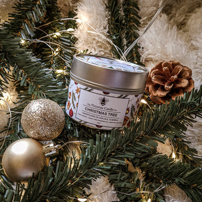 A cheerful arrangement showcasing The Victorian Candle Co.'s Christmas Tree - Scented Candle in a 100ml Tin 🎄, placed amidst lush evergreen branches, a pine cone, radiant warm white string lights, and two sparkling gold ornaments on a plush, white surface.