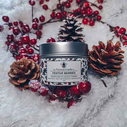 A Festive Berries - Scented Candle - 100ml Tin 🎅🏻 from The Victorian Candle Co. rests on a fluffy white surface, adorned with pine cones and red berries. Crafted from soy vegan-friendly wax, its cranberry scent envelops the air, evoking a warm holiday atmosphere.