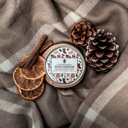A Spiced Clementine - Scented Candle - 100ml Tin from The Victorian Candle Co. sits on a plaid blanket, surrounded by dried orange slices, cinnamon sticks, and pinecones, creating a cozy, Christmas-scented atmosphere.