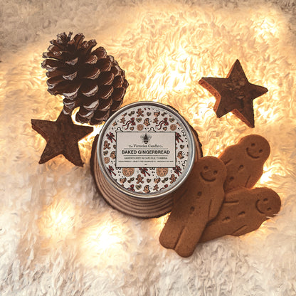 A cozy arrangement features a Baked Gingerbread - Scented Candle - 100ml Tin 🍪 from The Victorian Candle Co., casting a warm glow over two gingerbread cookies, star-shaped ornaments, and a pine cone on a fluffy white surface. This candle enriches the festive, wintry atmosphere with its inviting aroma.