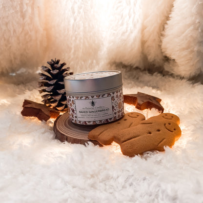 A 100ml tin of The Victorian Candle Co.'s Baked Gingerbread - Scented Candle 🍪 is perched on a wooden slice, surrounded by gingerbread cookies and a pinecone. This charming arrangement rests on a fluffy white surface, perfectly capturing a cozy, wintery atmosphere ideal for enjoying the delightful aroma of the candle.