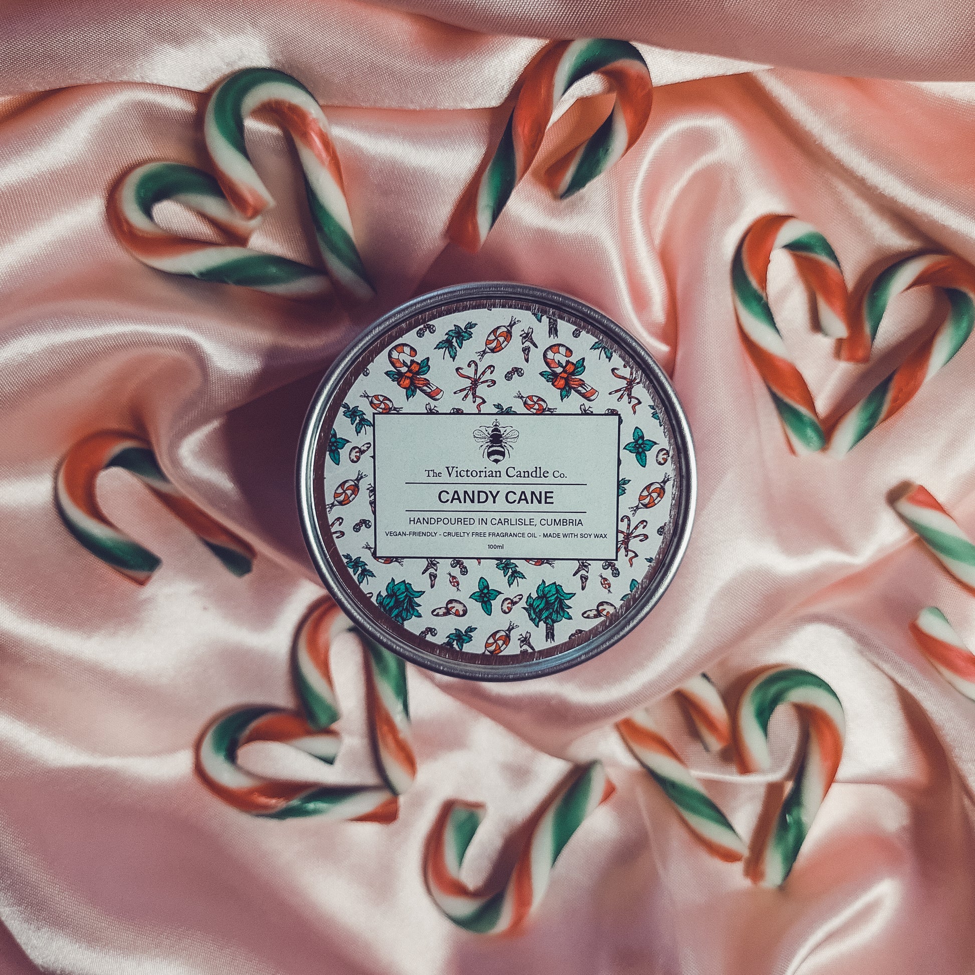 Set on pink satin fabric, the Candy Cane - Scented Candle - 100ml Tin ❄️ from The Victorian Candle Co. is surrounded by heart-shaped arrangements of candy canes. Crafted with soy wax, the candle's peppermint leaf fragrance is beautifully enhanced by the shiny texture of the fabric.