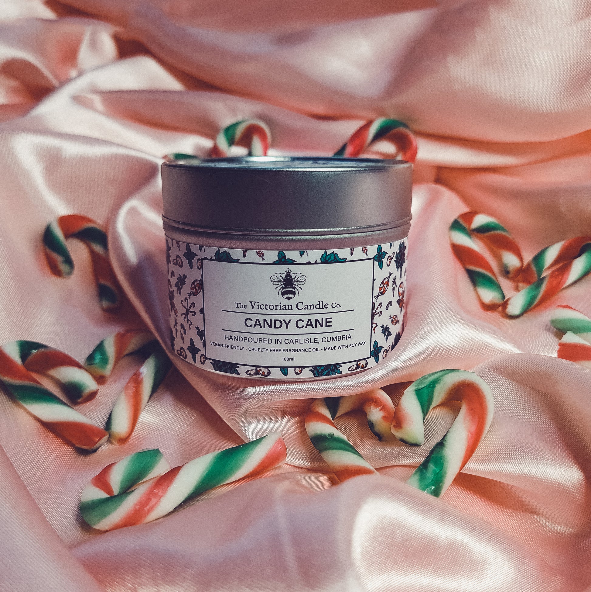 A 100ml tin of Candy Cane-scented candle from The Victorian Candle Co. is displayed on silky pink fabric, accompanied by small green and red candy canes. Infused with a peppermint leaf fragrance, the label indicates it's handmade in Carlisle, Cumbria.