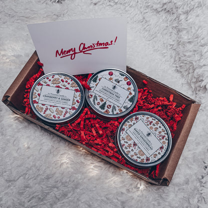 The Gift Wrapped - Christmas Trio Small Candle Box 🎁 from The Victorian Candle Co. includes three decorative tins labeled Cranberry & Ginger, Christmas Tree, and Snow Berries, each containing scented candles made from vegan-friendly wax. Resting on a fluffy white surface among red shredded paper is a card with the greeting "Merry Christmas!