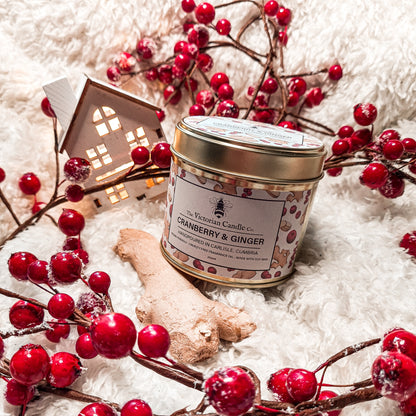 A (Limited Edition) Large Cranberry & Ginger - Scented Candle - 200ml Gold Tin 🌟 from The Victorian Candle Co. is elegantly displayed on a fluffy white surface, encircled by vibrant red berries, ginger root, and a quaint decorative wooden house with glowing windows. This festive fragranced arrangement exudes a cozy and inviting ambiance.