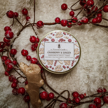 A limited edition Large Cranberry & Ginger scented candle from The Victorian Candle Co. rests amid red berries and a ginger slice on a soft, fluffy white surface. Its design is adorned with illustrations of cranberries and ginger, perfectly capturing the festive fragrance within its 200ml gold tin.