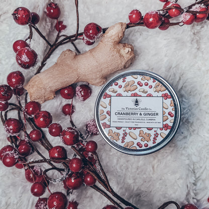 A 100ml tin of The Victorian Candle Co.'s Cranberry & Ginger Scented Candle rests on a plush surface, encircled by festive red berries and a slice of fresh ginger. Made with soy wax, the candle's lid highlights both the fragrance and decorative illustrations, making it ideal for the festive season.
