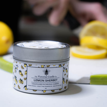 A Lemon Sherbet - Scented Candle - 100ml Tin from The Victorian Candle Co. with a vibrant yellow lemon design is displayed on the table. In the background, sliced lemons and a blurred hand enhance the invigorating citrus scent.