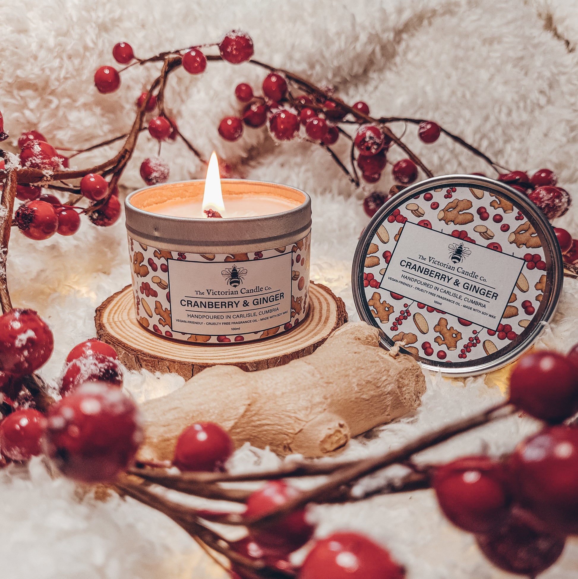 A Cranberry & Ginger - Scented Candle - 100ml Tin from The Victorian Candle Co. is nestled on a wooden coaster. Encircled by red berries, ginger root, and a gingerbread-design lid that complements it, the setting radiates the warmth of the festive season. Made with soy wax, this candle enhances the cozy atmosphere against a fluffy white backdrop.