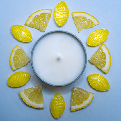 A lit Lemon Sherbet scented candle, presented in a 100ml tin from The Victorian Candle Co., is surrounded by alternating slices of lemon and yellow candies on a light blue background, arranged in a circular pattern, releasing a delightful citrus aroma.