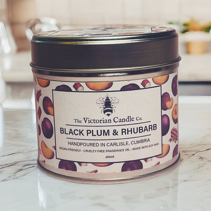 A (Large) Black Plum & Rhubarb Scented Candle in a 200ml tin by The Victorian Candle Co. rests on a reflective surface, releasing its fruity fragrance. The label, featuring fruit illustrations, proudly states that it is handcrafted in Carlisle from soy vegan-friendly wax.