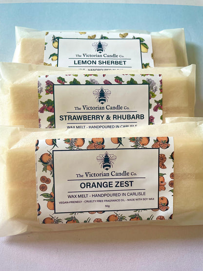 Three Blueberry Pancakes Wax Melt Snap Bars from The Victorian Candle Co. are stacked, beautifully enclosed in biodegradable packaging. Each scent is labeled with floral or fruit illustrations around the edges.