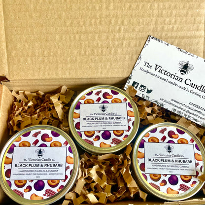 A Trio Box featuring three 100ml tins of Black Plum & Rhubarb scented candles, each adorned with decorative fruit labels, is presented in an open box lined with brown crinkle paper. These soy wax candles are accompanied by a business card from The Victorian Candle Co.