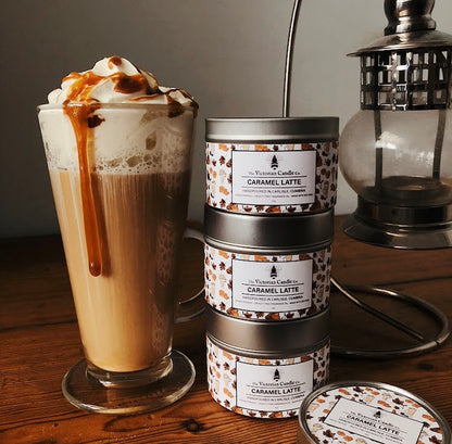 A tall glass of caramel latte, adorned with whipped cream and a caramel drizzle, rests beside three stacked tins of "Caramel Latte" scented candles by The Victorian Candle Co. on a wooden table, with a lantern setting the ambiance in the background.