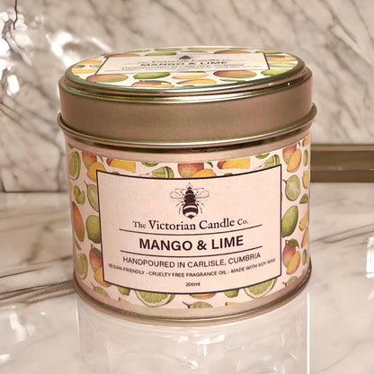 A (Large) Mango & Lime - Scented Candle - 200ml Tin from The Victorian Candle Co. sits on a reflective marble surface, its tin decorated with vibrant fruit illustrations. Crafted with soy wax, this vegan-friendly and cruelty-free candle brings an elegant touch to any space.