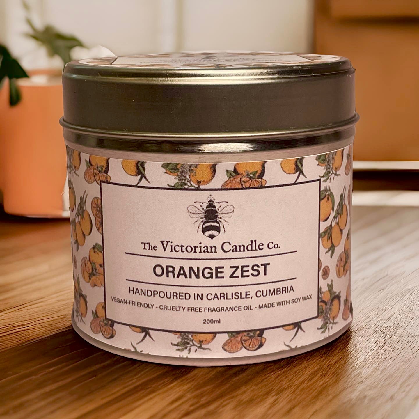 The (Large) Orange Zest - Scented Candle - 200ml Tin from The Victorian Candle Co. is set on a wooden surface, featuring a label with vibrant orange and leaf illustrations. Hand-poured in Carlisle, Cumbria, this vegan-friendly candle is made from soy wax to ensure a clean burn.