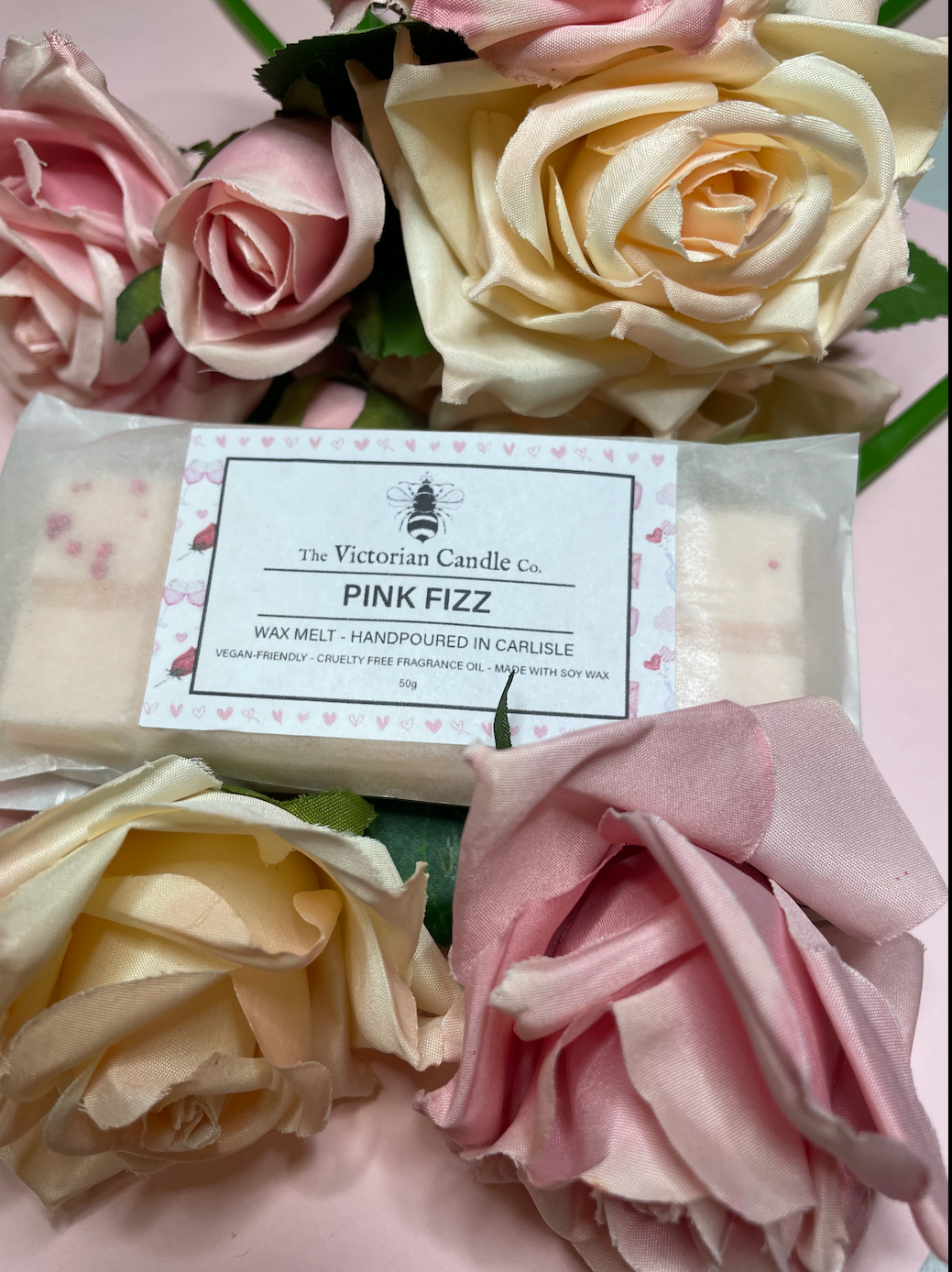 The Victorian Candle Co.s Limited Edition Pink Fizz Snap Bar, a Valentines Day wax melt nestled among cream and pink roses, features packaging stating handmade in Carlisle, vegan-friendly, and cruelty-free against a soft pink background.