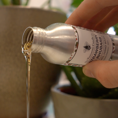 A hand pours scented oil from a silver bottle labeled Strawberry & Rhubarb Travel Hand Wash - 100ml by The Victorian Candle Co. into a container with green plants in the background, creating a serene and aromatic atmosphere reminiscent of a vegan-friendly strawberry and rhubarb experience.