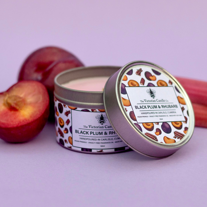 A 100ml tin of The Victorian Candle Co.'s Black Plum & Rhubarb Scented Candle rests on its side, with the lid slightly ajar, exposing the pink soy vegan-friendly wax inside. Plum and rhubarb elements are artfully displayed in the background against a gentle purple backdrop, accentuating its fruity fragrance.