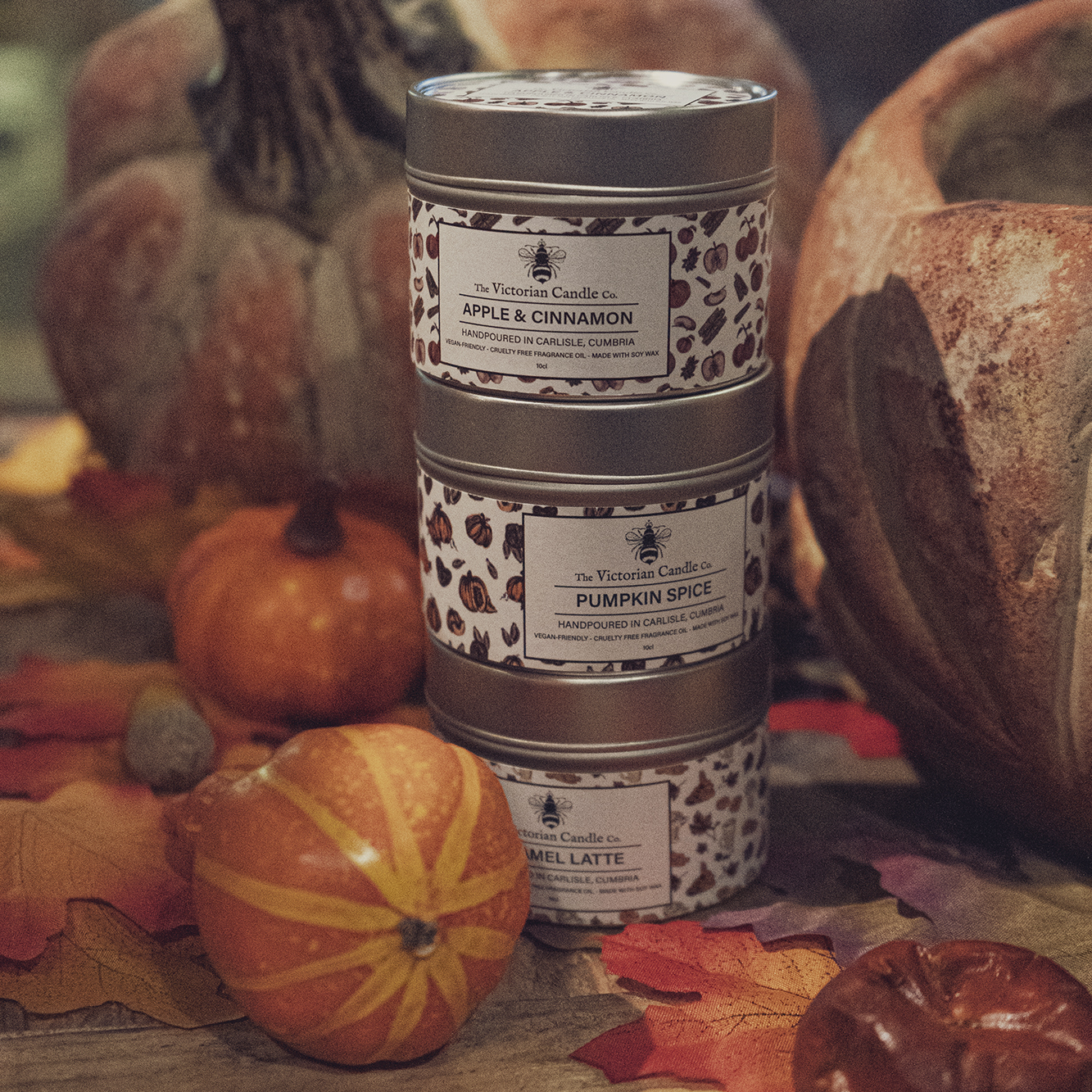 A stack of three decorative soy wax candle tins labeled Apple & Cinnamon, Pumpkin Spice, and Caramel Latte is surrounded by pumpkins and autumn leaves. The Victorian Candle Co.'s Autumn Trio 🍂 scented candles beautifully evoke a cozy fall atmosphere.