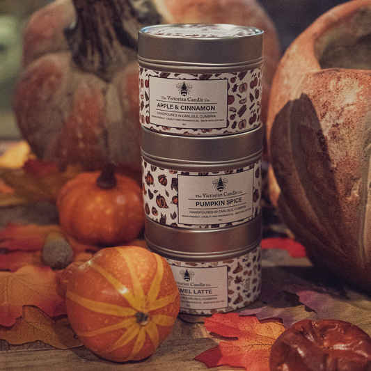 A stack of three decorative soy wax candle tins labeled Apple & Cinnamon, Pumpkin Spice, and Caramel Latte is surrounded by pumpkins and autumn leaves. The Victorian Candle Co.'s Autumn Trio 🍂 scented candles beautifully evoke a cozy fall atmosphere.